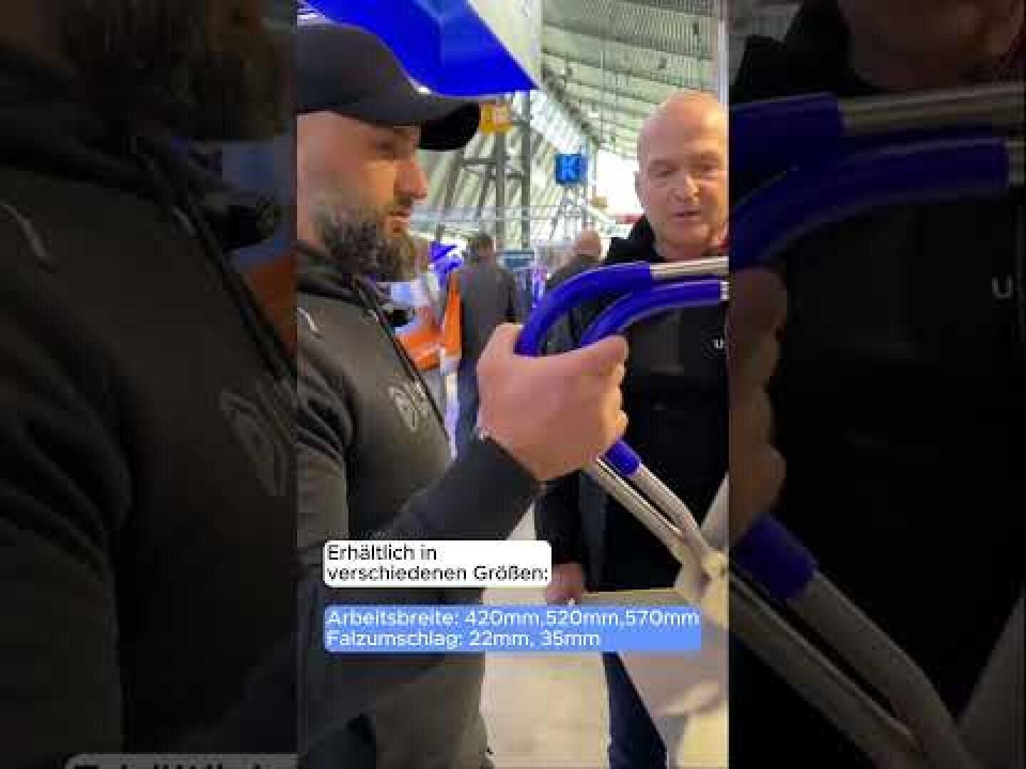 Zwei Männer stehen in einem Haus, einer hält einen blau-silbernen Klappartikel. Ein Schild in deutscher Sprache mit Maßangaben ist zu sehen. Im Hintergrund sind Regale und Produkte zu sehen, was auf ein Geschäft hindeutet.