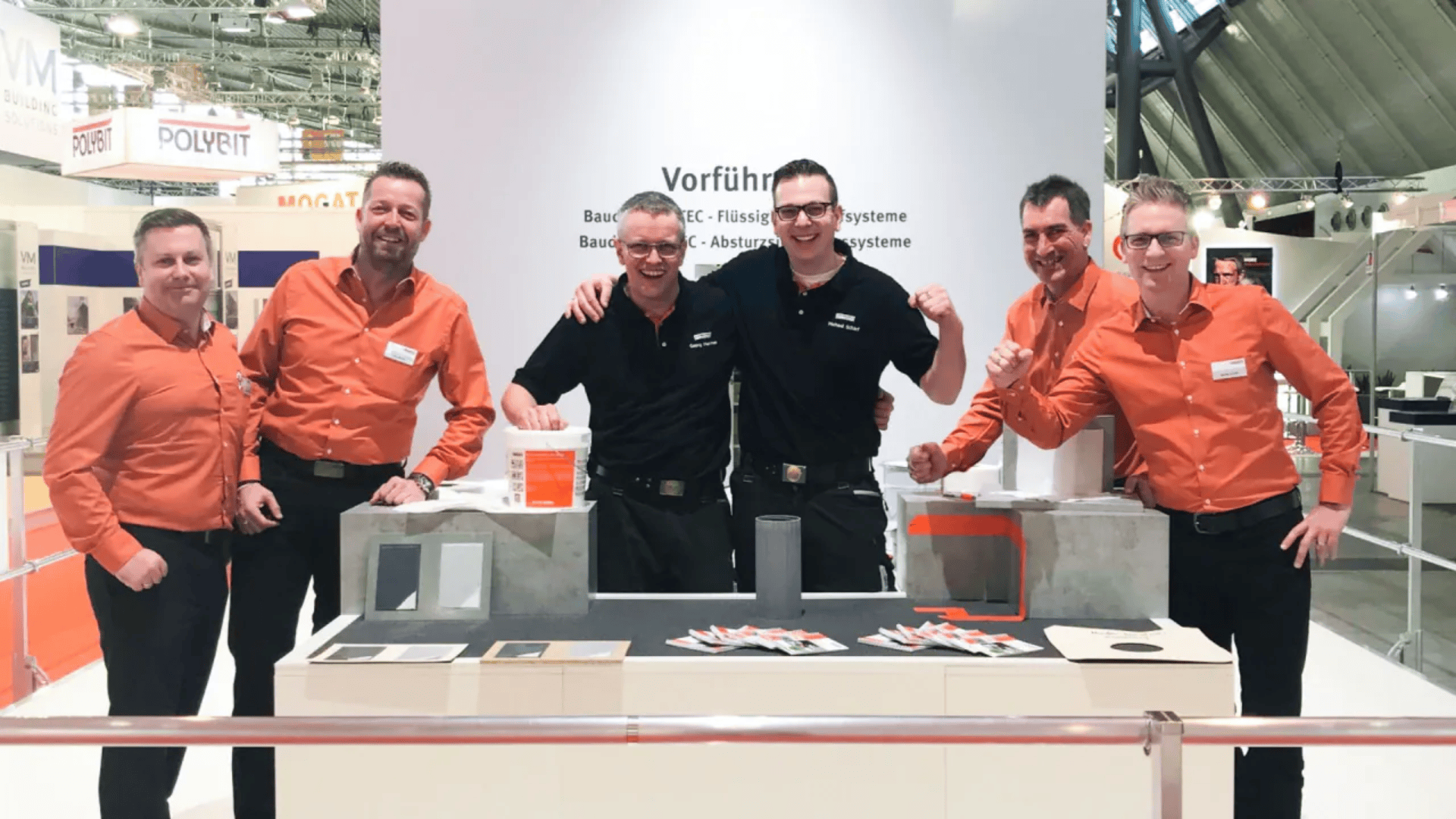 Fünf Männer stehen lächelnd hinter einem Ausstellungsstand. Drei tragen schwarze, zwei orangefarbene Hemden. Am Stand werden Baumaterialien und Muster präsentiert. Auf weißem Hintergrund ist deutscher Text zu sehen. Die Szenerie scheint eine Messe zu sein.