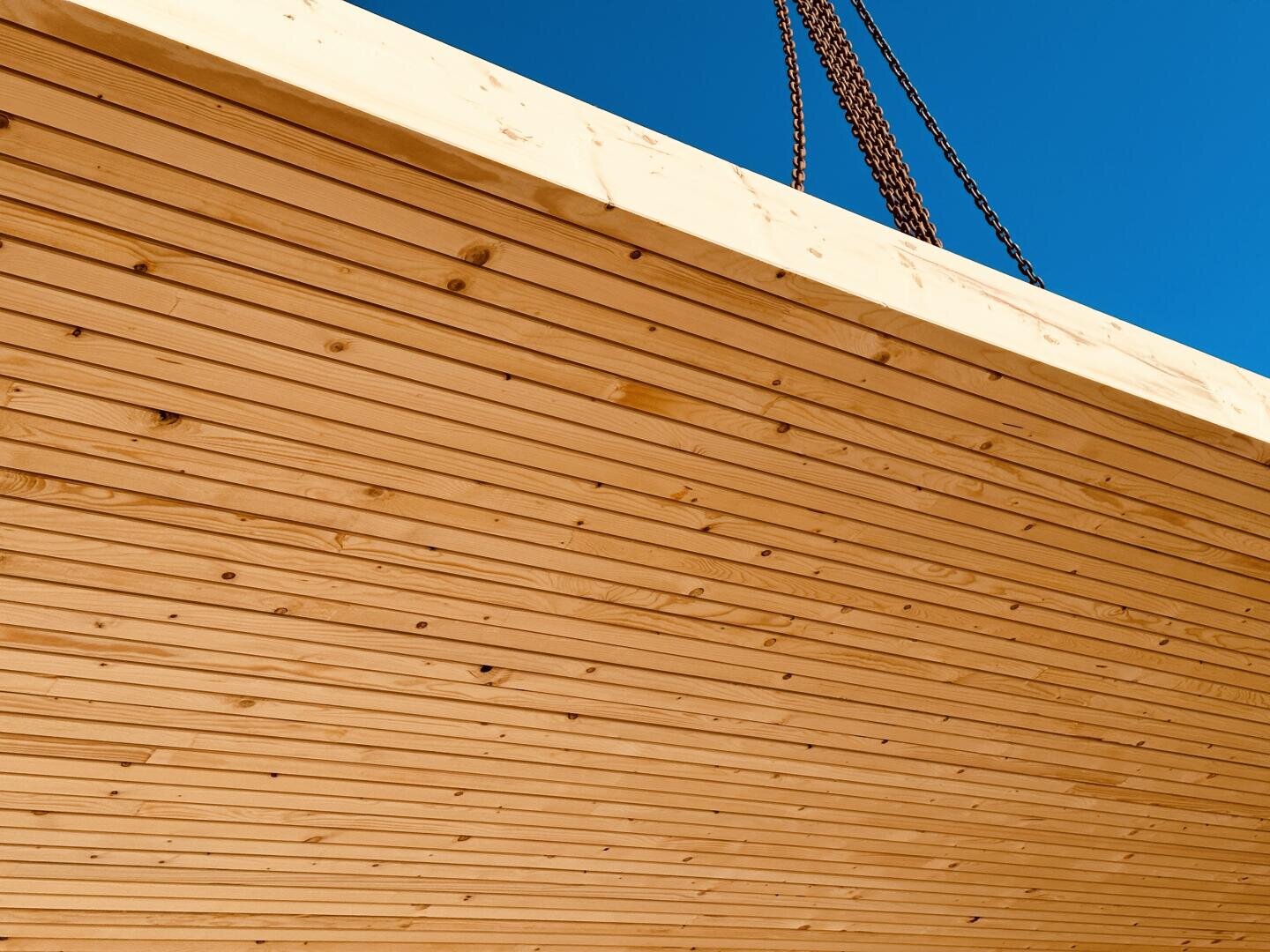 Large wooden beam with visible planks and grain, lifted by two thick reddish-brown chains against a clear blue sky.