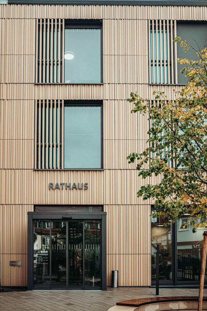 Modernes Gebäude mit vertikalen Holzlatten, großen Fenstern und einem gläsernen Eingang. Das Wort Rathaus steht über der Tür. Im Vordergrund rechts steht ein Baum mit grünen Blättern.