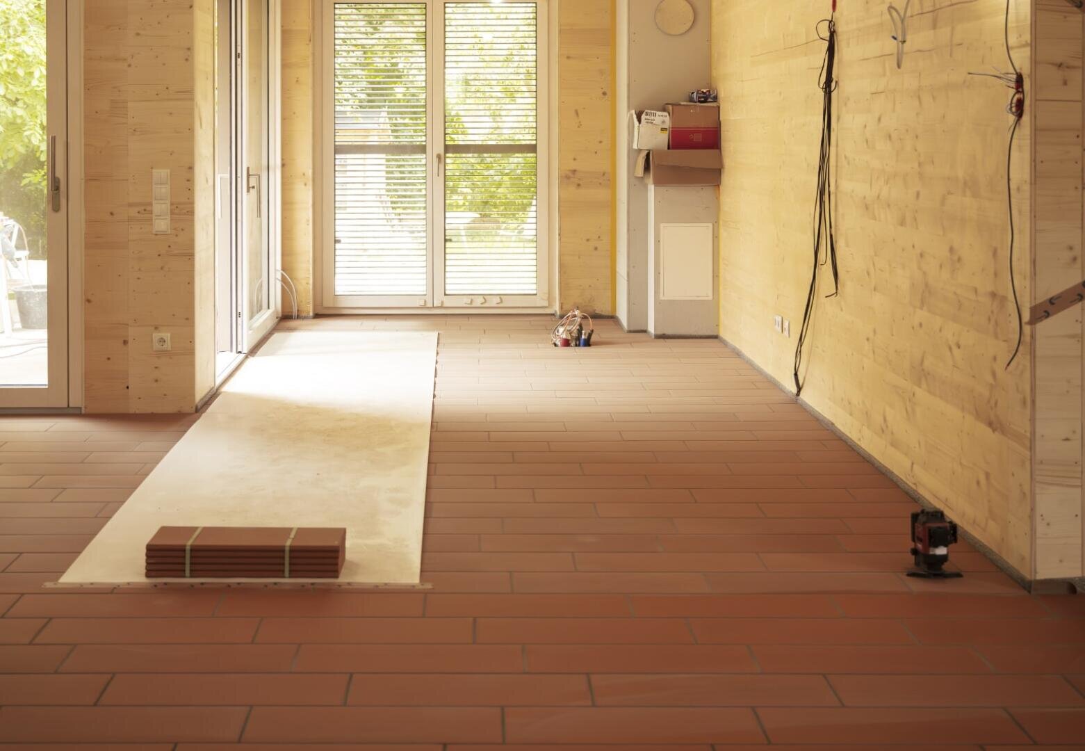 Ein Raum mit hellen holzgetäfelten Wänden und großen Fenstern wird gerade renoviert. Ein Teil des Fußbodens ist mit neuen braunen Fliesen ausgelegt, auf denen ein Stapel Fliesen und Werkzeuge zu sehen ist. Elektrische Leitungen hängen von den Wänden herab.