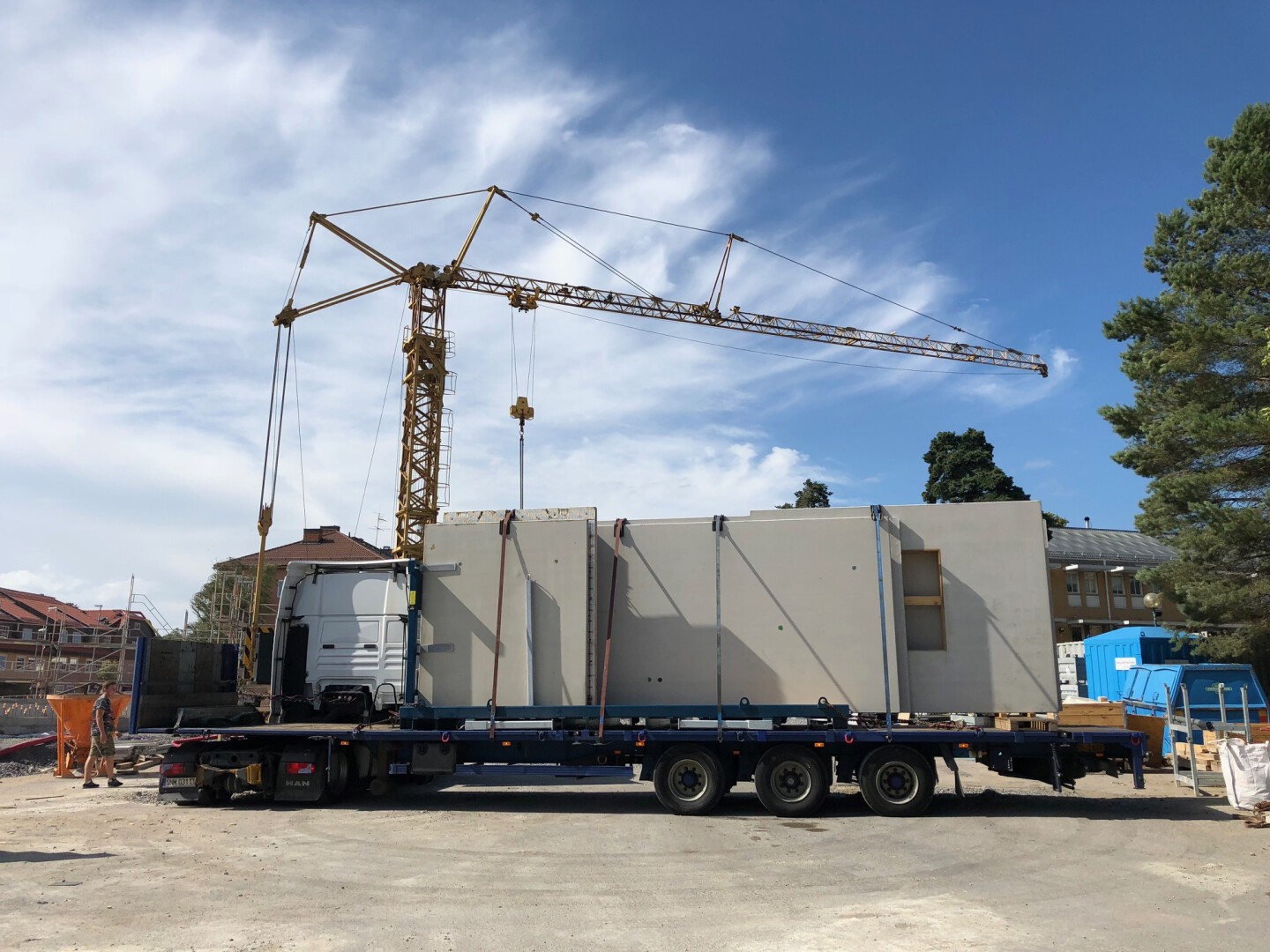 Ein Baukran hebt auf einer Baustelle ein großes vorgefertigtes Wandelement von einem Tieflader. Arbeiter in orangefarbener Schutzausrüstung stehen in der Nähe unter einem teilweise bewölkten blauen Himmel.