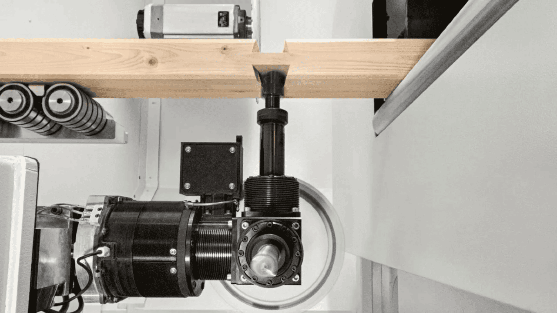 A close-up view of a CNC woodworking machine carving a precise rectangular notch into a wooden beam, with various machine components and tools visible around the cutting area.