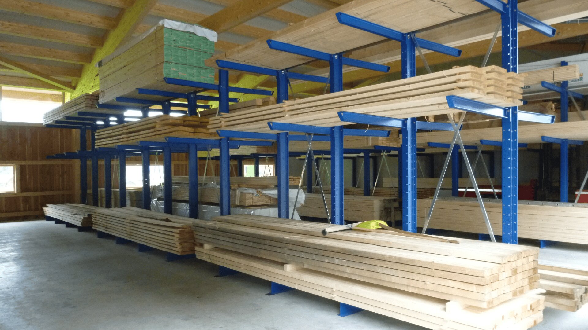 In einem Lagerhaus mit Betonboden und Holzbalkendecke stapeln sich Holzbretter auf blauen Metallgestellen. Das Holz ist ordentlich in Reihen in unterschiedlichen Höhen angeordnet.