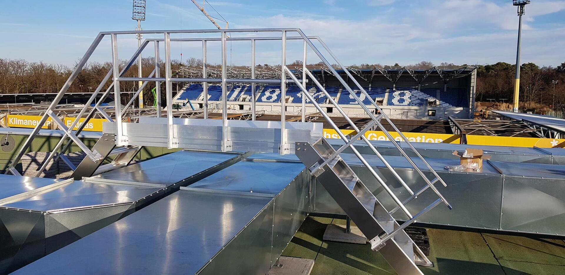 Ein Metalldachsteg mit Geländer und Treppe ist über Luftschächten installiert und bietet einen Blick auf ein Sportstadion mit blauen Sitzen und gelber Werbung. Im Hintergrund sind Bäume und Flutlichter zu sehen.