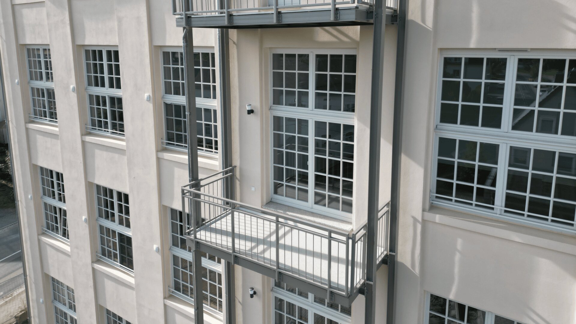 Ein beiges Gebäude mit großen Gitterfenstern und einem kleinen Balkon mit Metallgeländer. Der Balkon ist leer, und die Außenfassade des Gebäudes wirkt schlicht und modern.