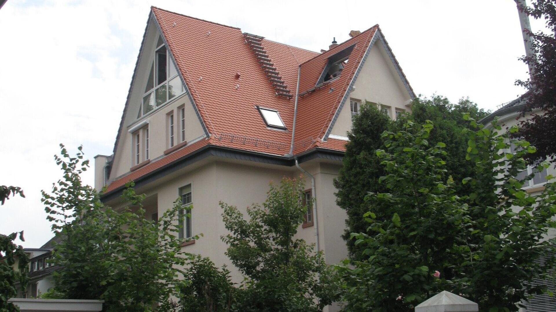 A beige house with a steep triangular red-tiled roof, multiple large windows, and skylights, surrounded by lush green trees and shrubs on a cloudy day.