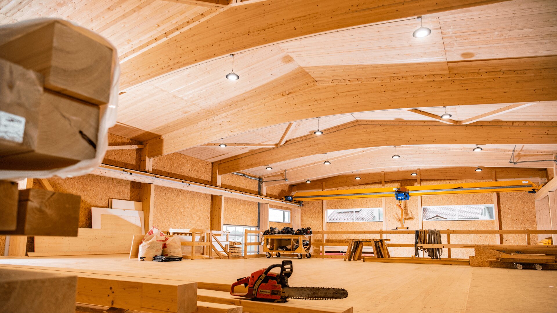 Innenraum einer geräumigen Holzwerkstatt mit gewölbten Decken, freiliegenden Balken und großen Fenstern. Eine Kettensäge sowie verschiedene Holzbearbeitungsmaterialien und -werkzeuge liegen auf dem Boden und den Werkbänken verteilt. Helle Deckenleuchten erhellen den Raum.