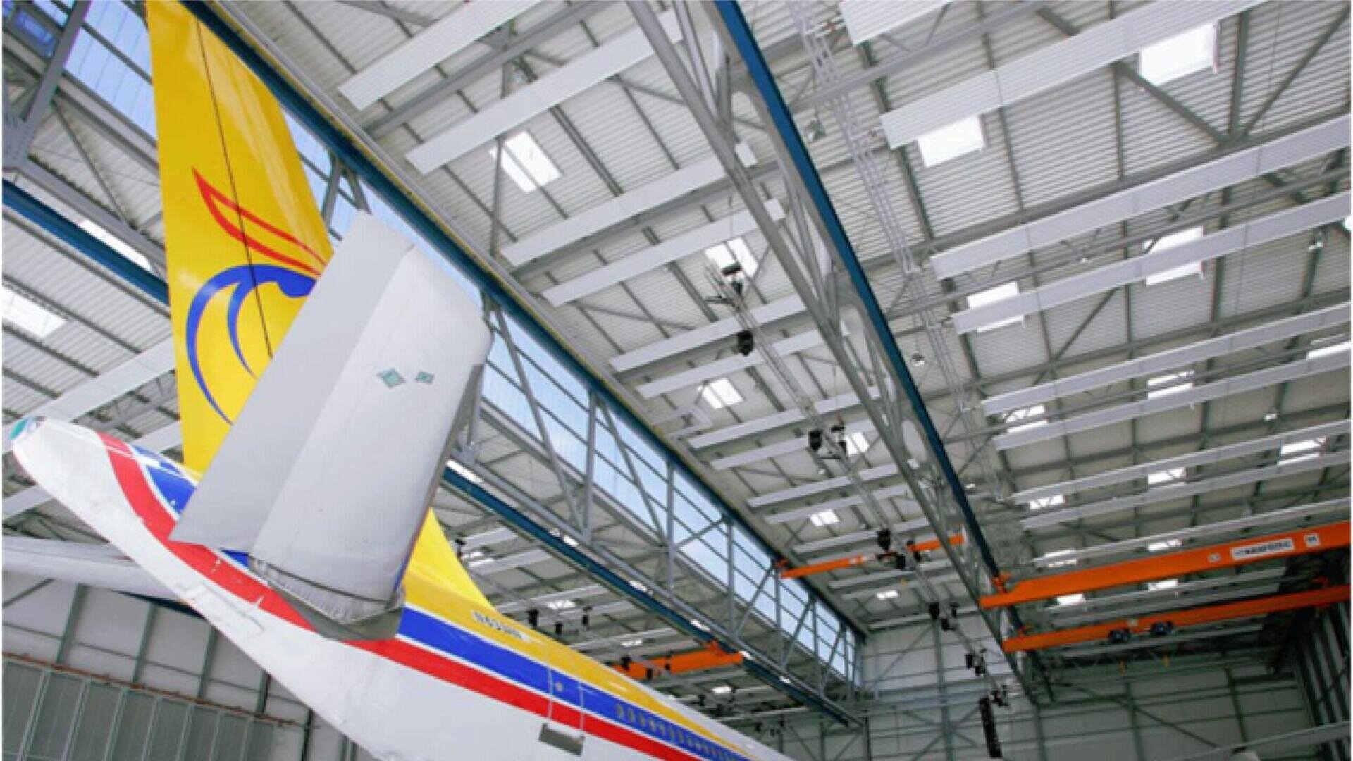 The tail section of a colorful airplane with a yellow, red, and blue design is inside a large, modern aircraft hangar with a high ceiling and visible metal beams.