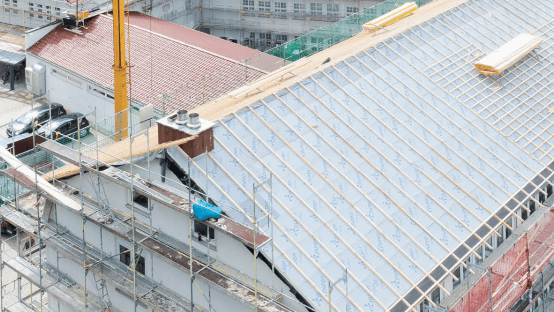Ein teilweise fertiggestelltes Gebäude mit Gerüst, einem schrägen, mit Dämmmaterial bedeckten Dach, freiliegenden Holzbalken und einem gelben Kran daneben. Auf dem Dach und in der Nähe sind Baumaterialien sichtbar.