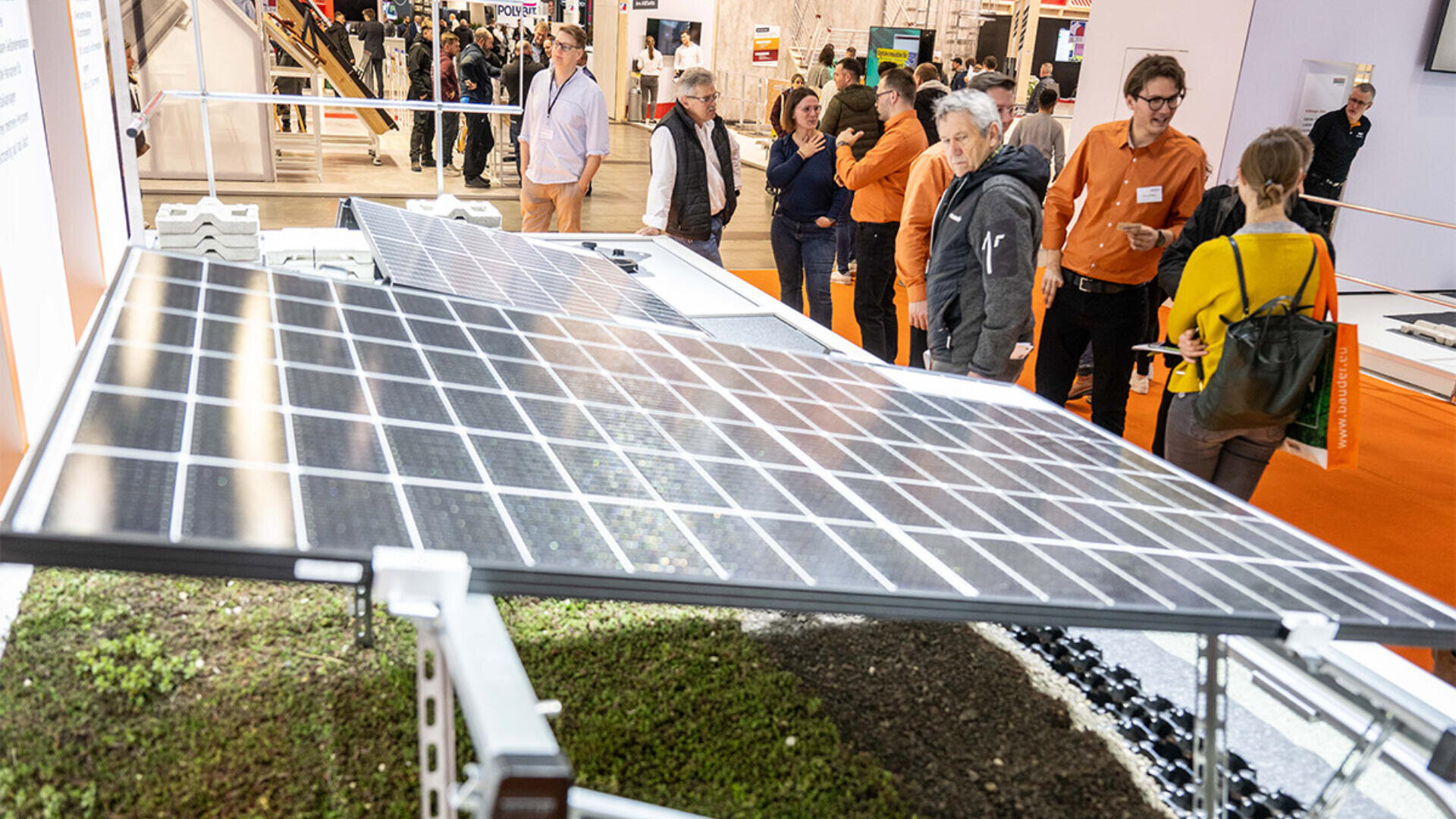 Eine Gruppe von Menschen betrachtet bei einer Indoor-Ausstellung eine große Solarpanel-Ausstellung über einem Beet mit grünen Pflanzen, im Hintergrund befinden sich Informationsstände und ein orangefarbener Teppich.