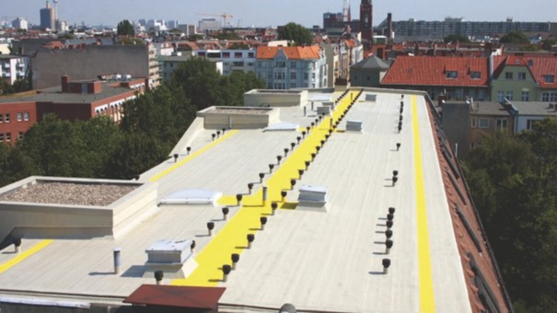Ein flaches, helles Dach mit gelben Gehwegen verläuft über mehrere miteinander verbundene Gebäude, umgeben von Bäumen und städtischen Wohnstrukturen unter einem klaren Himmel.