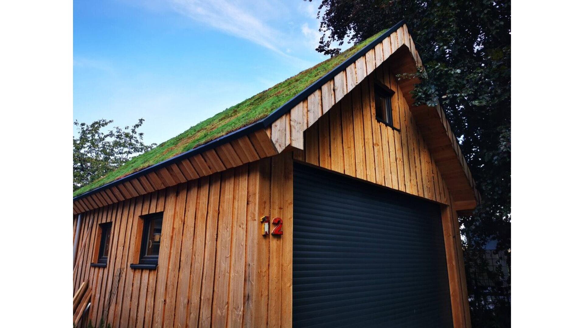 A wooden building with a grass-covered sloped roof, a black roll-up garage door, and small windows, set beside leafy trees under a partly cloudy blue sky. The number 12 is mounted near the door.