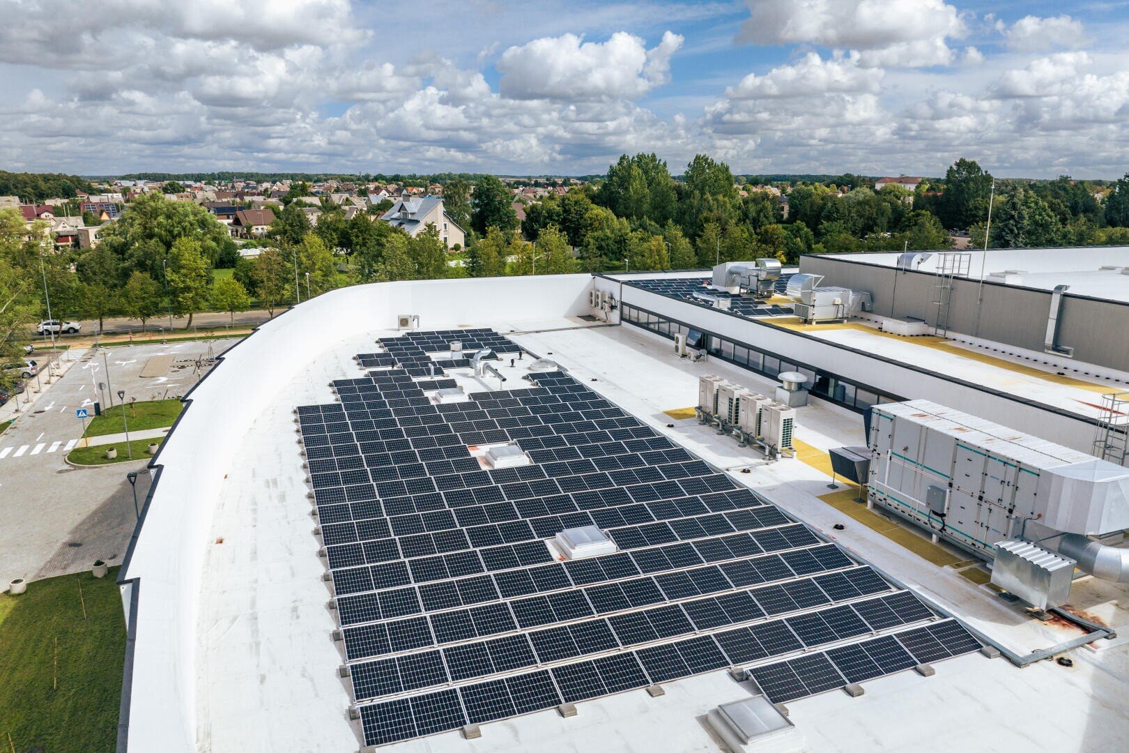 Aerial view of a large, flat, white rooftop with numerous solar panels installed, surrounded by trees and residential houses under a partly cloudy sky.