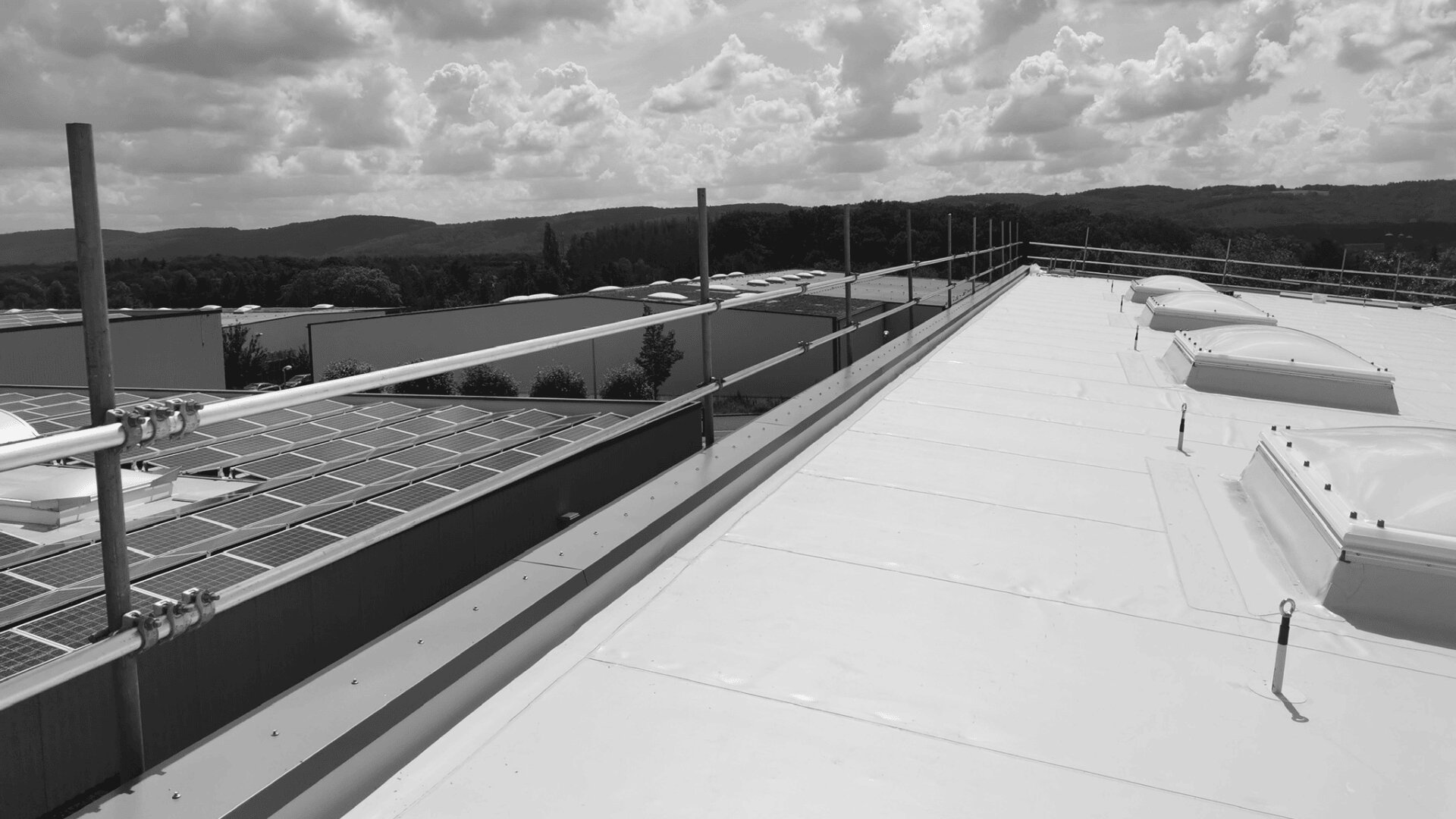 Ein flaches weißes Dach mit Sicherheitsgeländer und Oberlichtern neben Reihen von Solarmodulen. Im Hintergrund sind Industriegebäude und baumbedeckte Hügel unter einem bewölkten Himmel zu sehen.