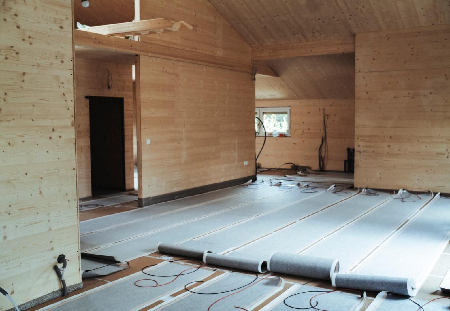 Ein Raum mit unfertigen Holzwänden und -decken befindet sich im Bau; auf dem Boden liegen Stoffrollen und freiliegende elektrische Heizkabel, und ein kleines Fenster lässt Tageslicht herein.