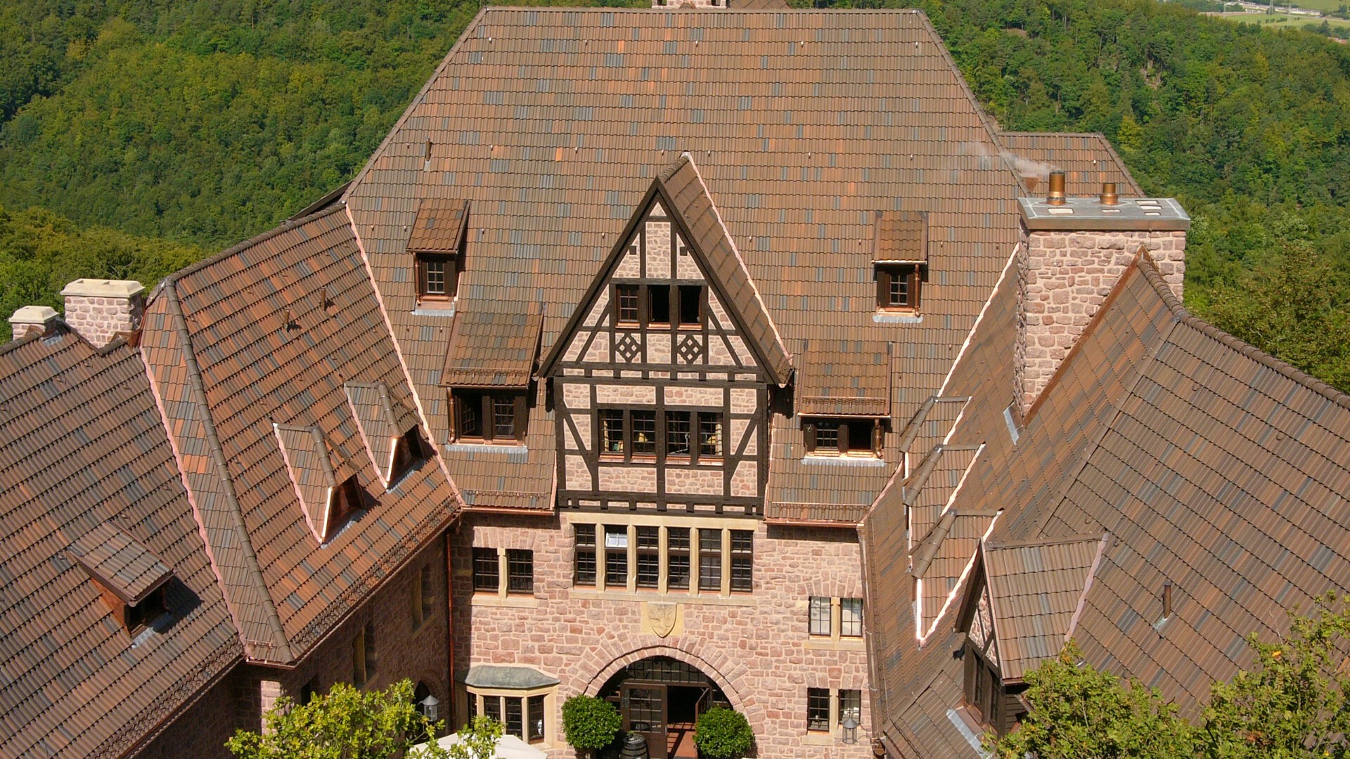 Großes Steingebäude mit kunstvollen braunen Dachziegeln, Fachwerkdetails, Bogenfenstern und einem zentralen Eingang, umgeben von üppigen grünen Bäumen und Hügeln im Hintergrund.