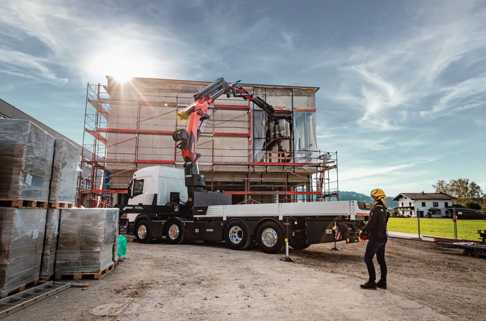 A worker in protective gear operates a crane mounted on a flatbed truck on a construction site next to a scaffolded building under a blue sky with clouds and sunshine.