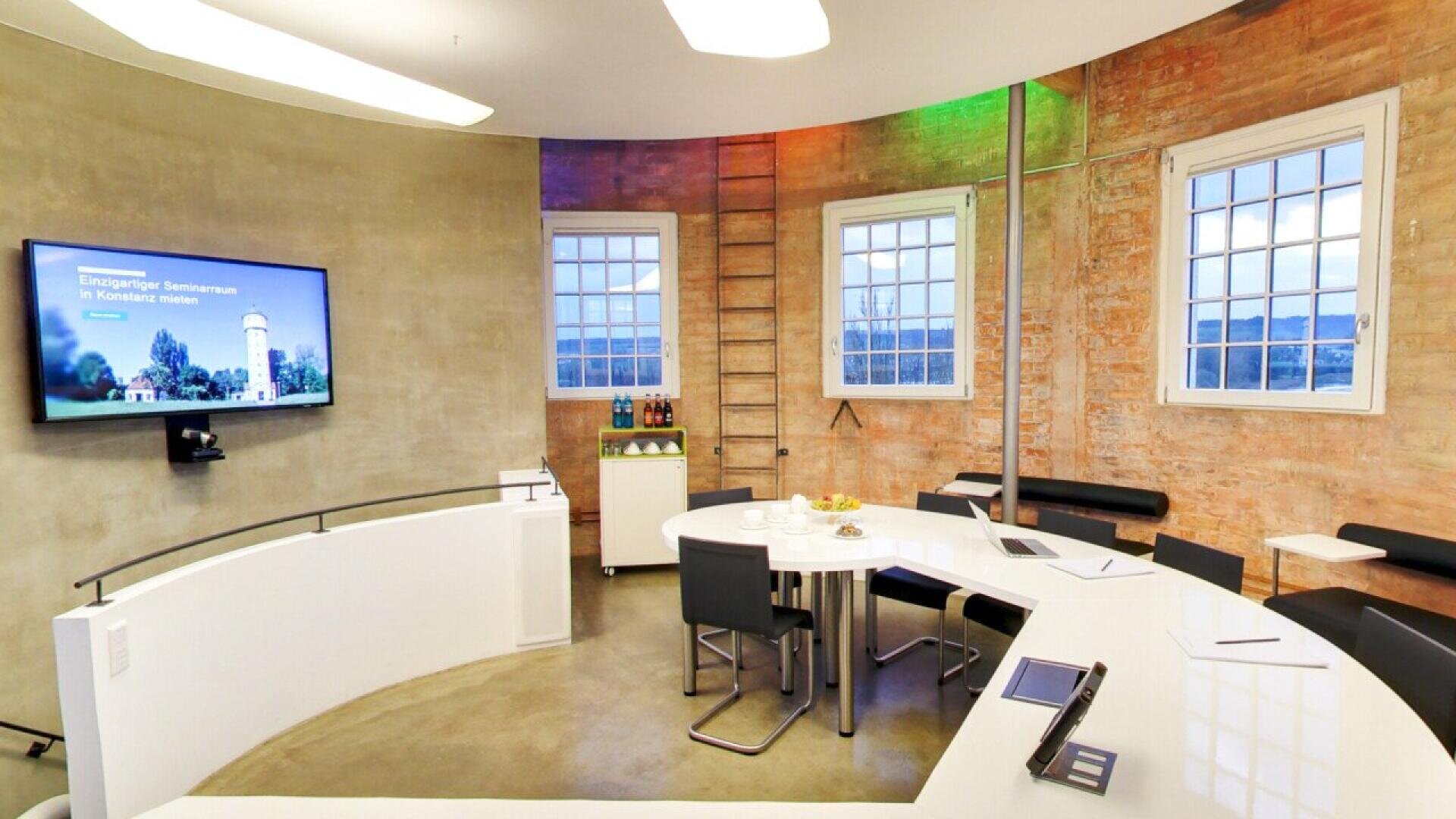 Modern meeting room with curved white tables, black chairs, exposed brick walls, large windows, a wall-mounted TV screen, a wooden ladder, and refreshments on a side table. The room is brightly lit and organized.
