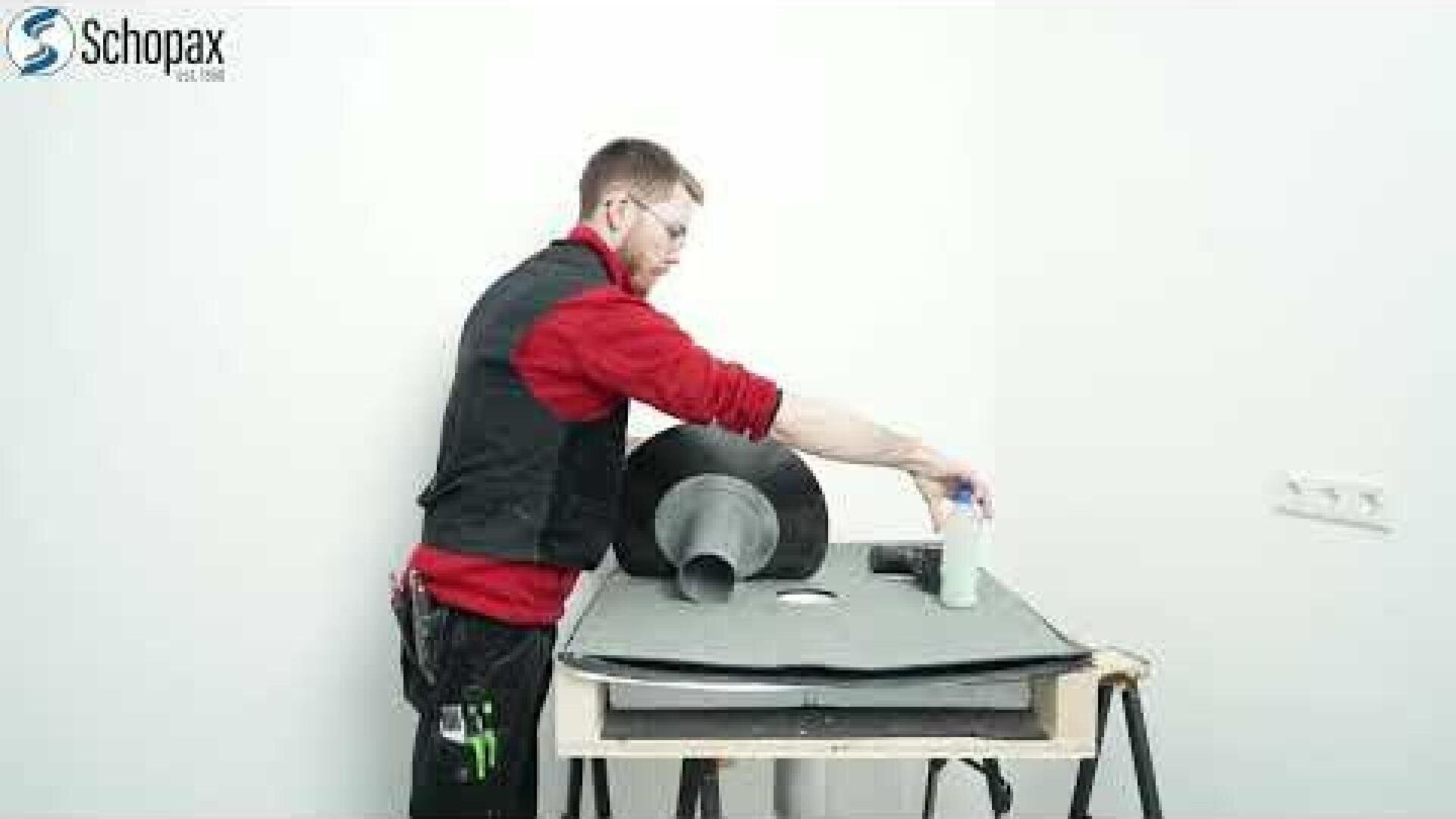 A person in a red shirt and black vest is working in a white room at a table with plumbing materials, including pipes and fittings. The Schopax logo can be seen in the top left corner.