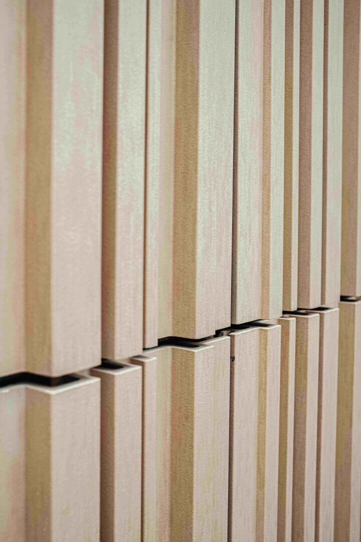 Eine Nahaufnahme einer modernen Wand mit vertikalen, rechteckigen beigen Paneelen, die in einem sich wiederholenden Muster angeordnet sind und ein strukturiertes und geometrisches Aussehen erzeugen.