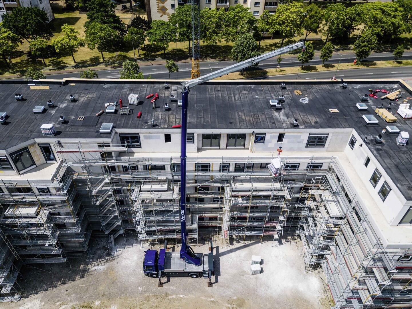 Luftaufnahme einer Baustelle mit Gerüsten um ein großes, U-förmiges Gebäude. Ein Kranwagen hebt Materialien auf das Dach, und mehrere Arbeiter sind auf dem Gerüst und auf dem Dach zu sehen. Im Hintergrund sind Bäume und eine Straße zu sehen.