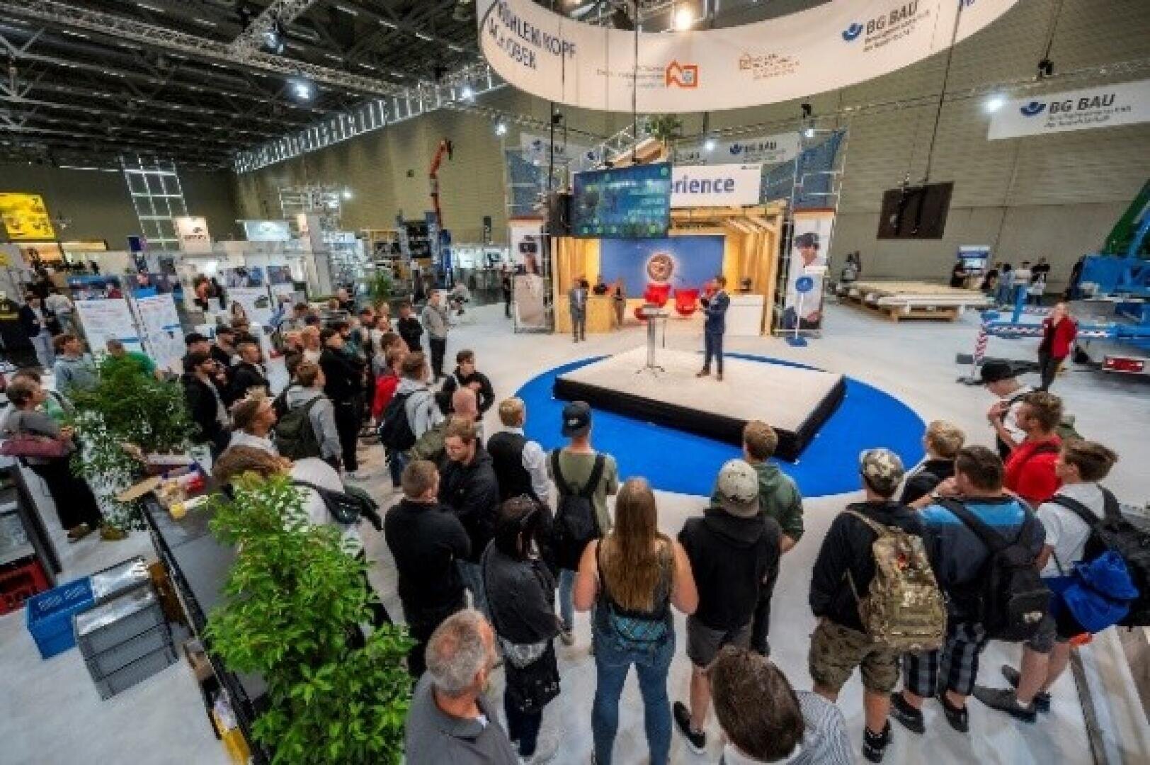 Eine Menschenmenge versammelt sich um eine kleine Bühne auf einer überdachten Fachmesse oder Expo und hört einem Redner zu. In der geräumigen, gut beleuchteten Messehalle sind verschiedene Stände, Banner und Ausrüstungsdisplays zu sehen.