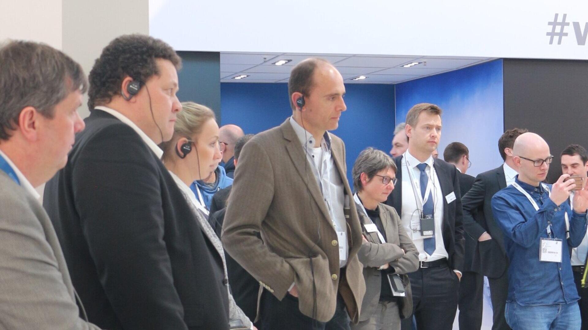 A group of professionally dressed people, some wearing headsets, stand attentively in a modern indoor setting, possibly at a conference or presentation.