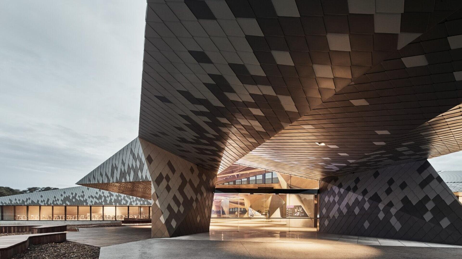 A modern building with angular, geometric overhangs and a patterned metal facade, illuminated by warm lights, creating dramatic shadows and reflections on the ground.