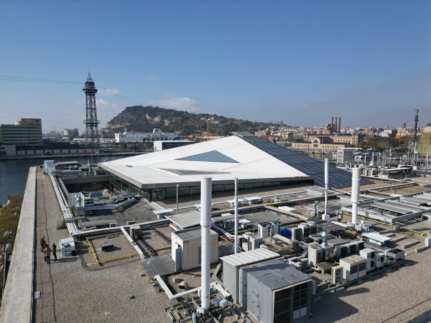 Ein modernes Gebäude mit weißem, eckigem Dach und sichtbaren Dachgeräten in der Nähe eines Hafens. Im Hintergrund sind ein grüner Hügel und städtische Gebäude unter einem klaren blauen Himmel zu sehen.