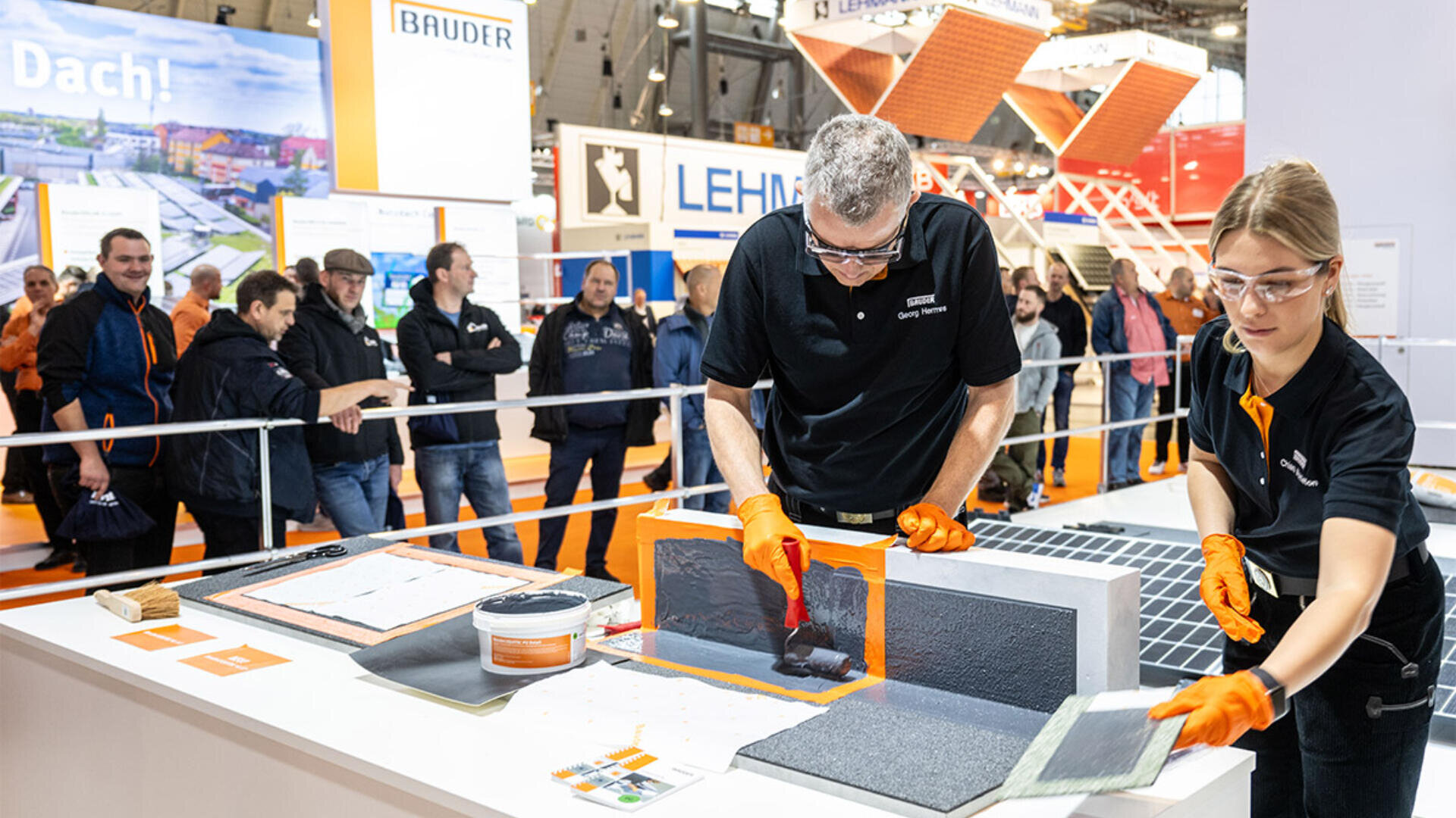 Zwei Personen in schwarzen Hemden, orangefarbenen Handschuhen und Schutzbrillen demonstrieren mit Werkzeugen und Materialien auf einem Tisch Dachdecker- oder Bautechniken, während eine Menschenmenge bei einer Indoor-Ausstellung zusieht.