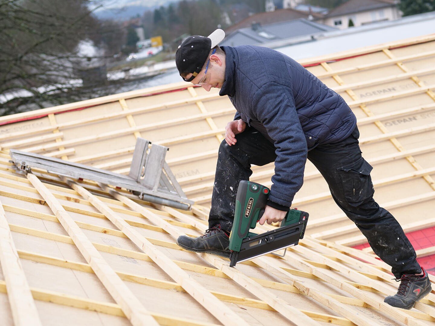 Eine Person in Marinejacke, schwarzer Hose, Mütze und Schutzbrille befestigt mit einem Nagelgerät Holzbretter auf einem im Bau befindlichen Schrägdach. In der Nähe steht eine Leiter, im Hintergrund sind Häuser zu sehen.
