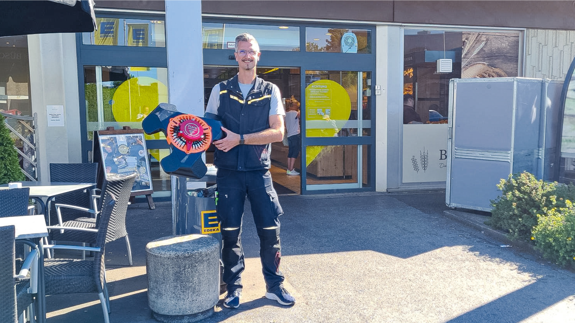 Ein lächelnder Mann in dunklem Outfit steht vor einem Gebäude und hält eine große bunte Pappfigur in Form einer Blume in der Hand. In der Nähe stehen Tische und Stühle, und das Sonnenlicht wirft Schatten auf den Boden.