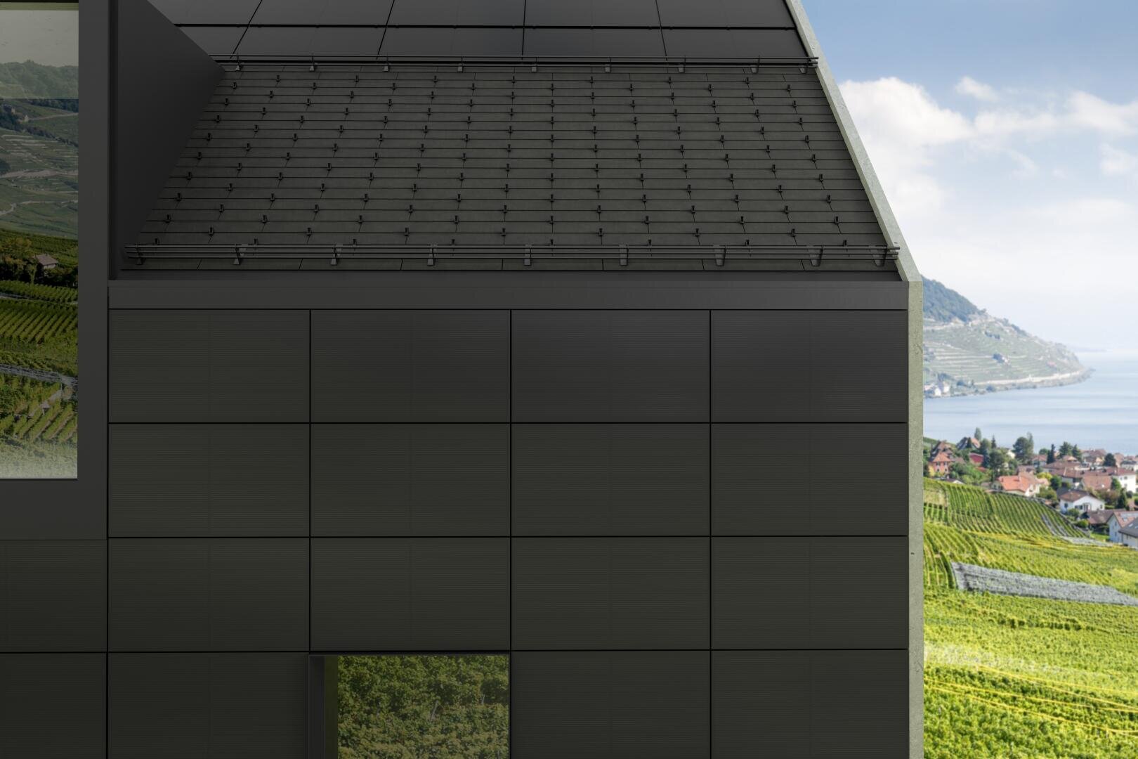 Modern black building with solar panels on the roof, large tiles on the outside and a window reflecting green vineyards. In the background are rolling hills, vineyards, houses and a blue lake under a partly cloudy sky.