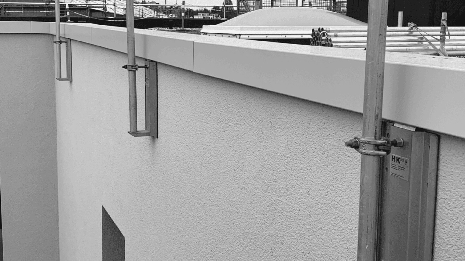 A close-up view of a building’s exterior wall with scaffolding and construction materials on the rooftop, shown in black and white. The wall has a textured finish and there are metal brackets attached.
