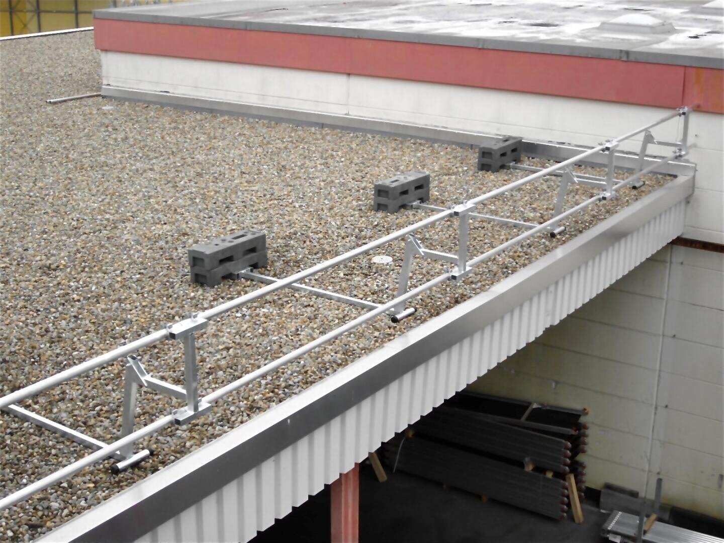 A metal safety railing is installed along the edge of a flat, gravel-covered roof that is anchored with concrete blocks and overhangs the side of a building.