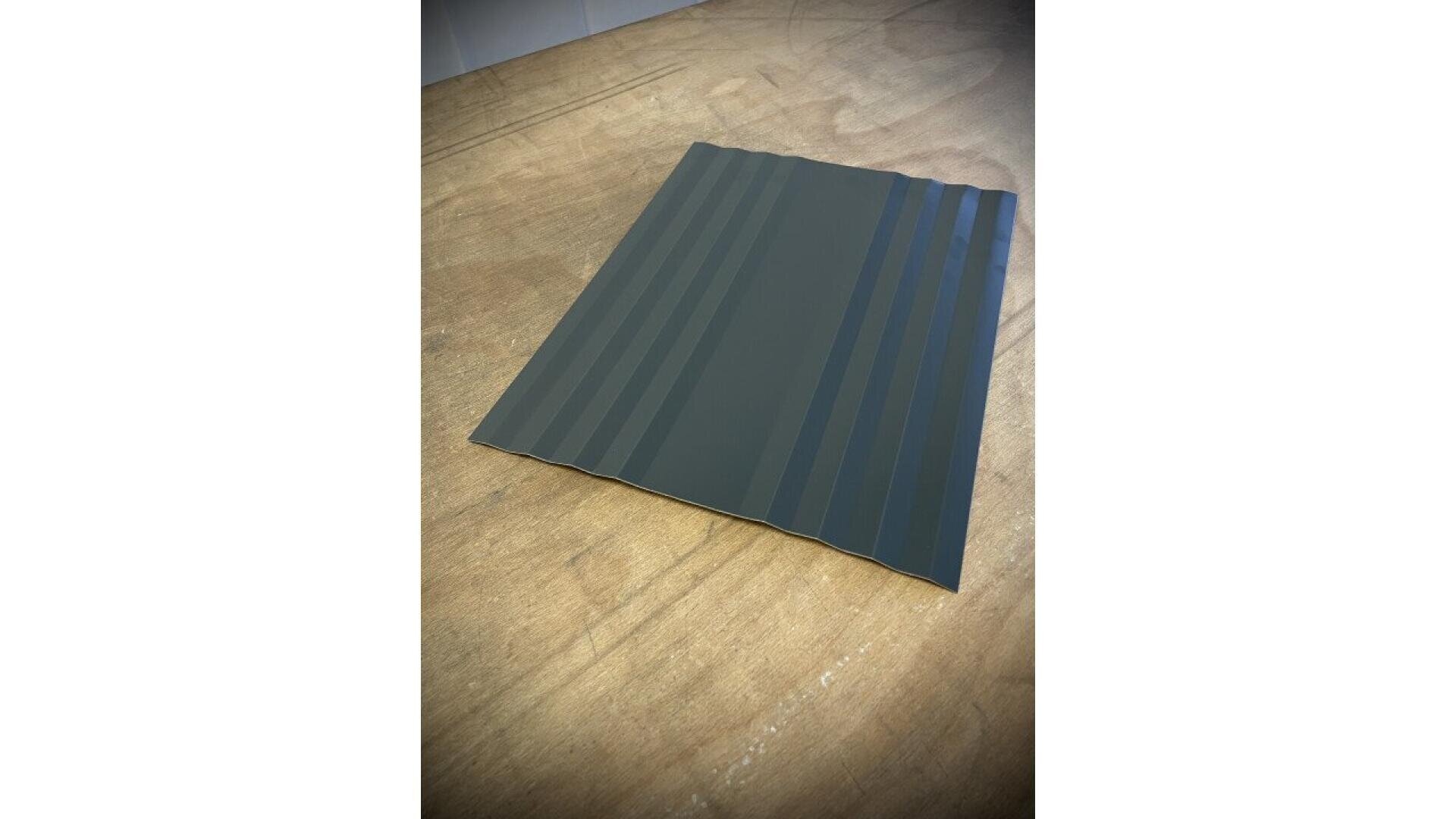 A rectangular sheet of dark gray corrugated metal with alternating wide and narrow ridges, placed on a light brown wooden surface.