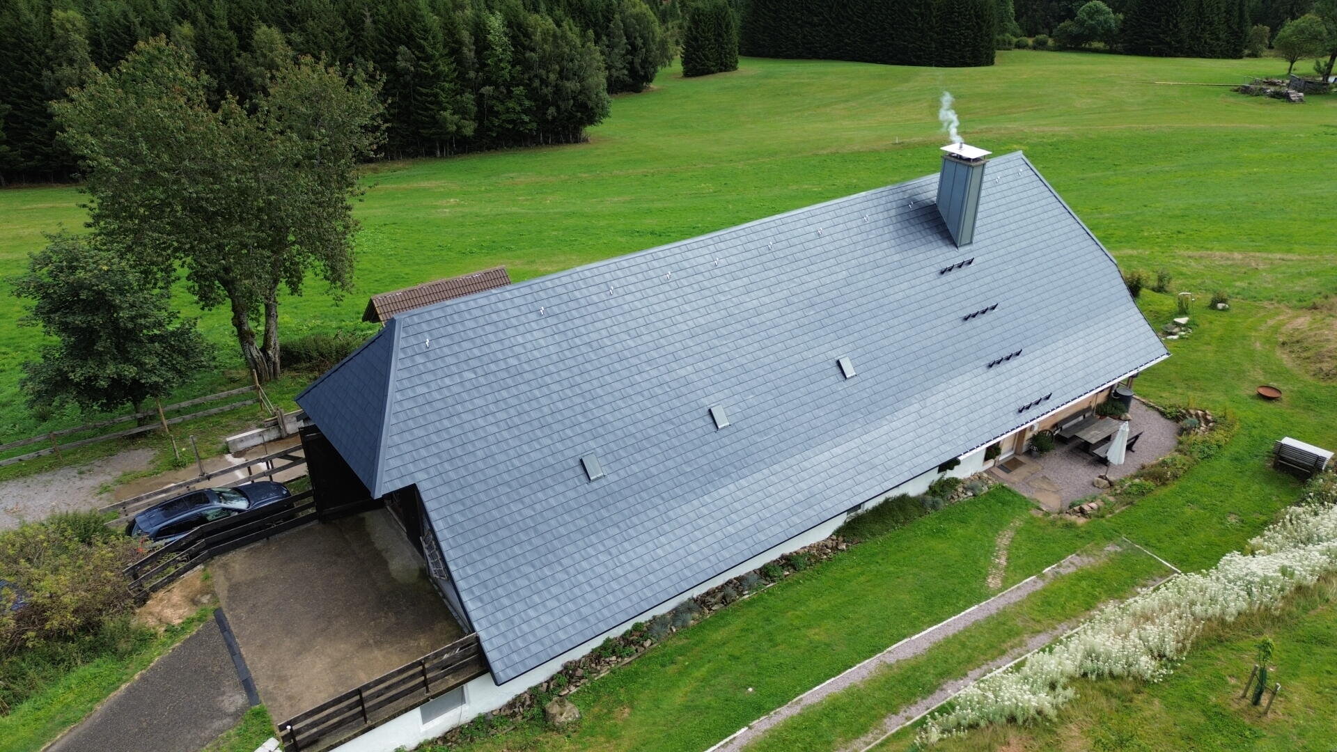 Aerial view of a long, gray-roofed building with smoke rising from a chimney, surrounded by green grass, trees, and a gravel driveway where a car is parked.