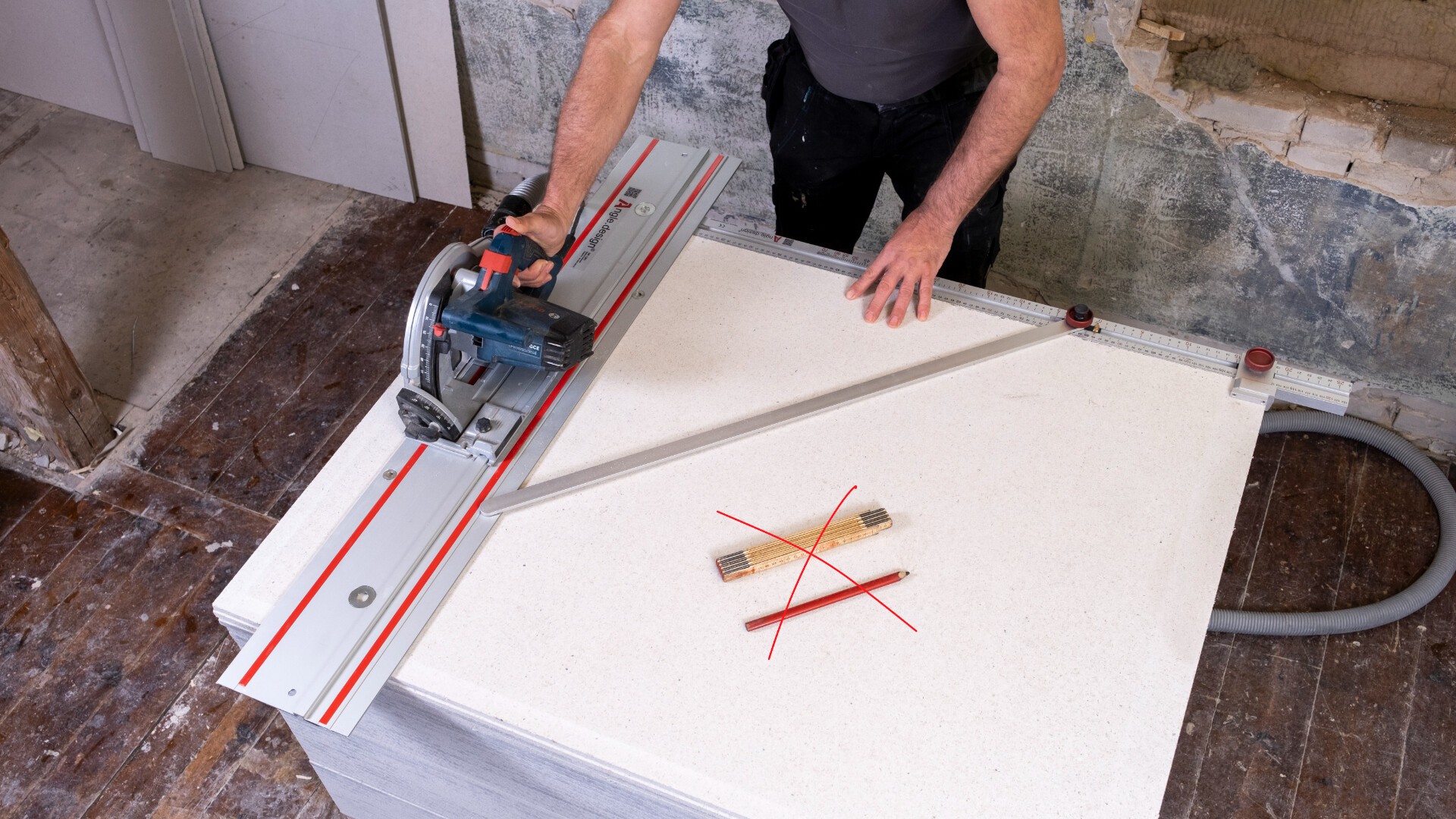 A person uses a circular saw with a guide rail to cut a large white board. On the board, there is a measuring tool and pencils crossed in an X shape, marked with a red X. The workspace has unfinished walls and a wooden floor.