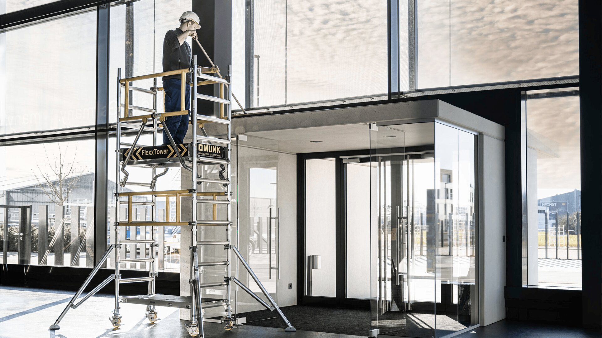 Ein Arbeiter mit Helm steht auf einem mobilen Gerüst und reinigt hohe Glasfenster in einem modernen Gebäude mit großen Glastüren und einfallendem Tageslicht.