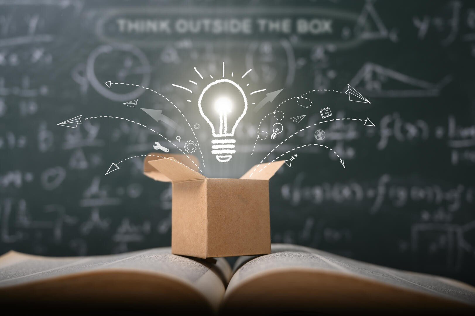 Eine leuchtende Glühbirne taucht aus einer Pappschachtel über einem aufgeschlagenen Buch auf, das mit Pfeilen, Papierfliegern und Symbolen bekritzelt ist, vor einer Kreidetafel mit Formeln und der Aufschrift THINK OUTSIDE THE BOX.