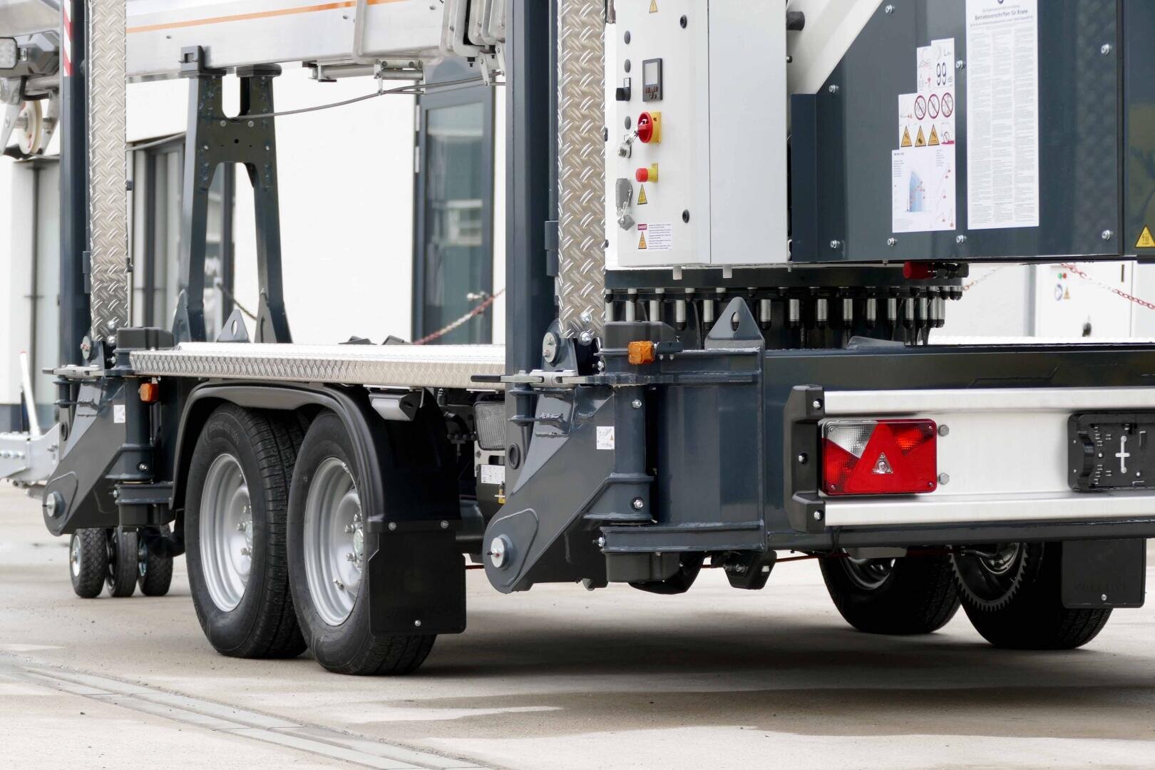 Close-up of the rear wheels and lower part of a large industrial machine or vehicle with metal frame, control panels, warning signs and exposed machine parts.