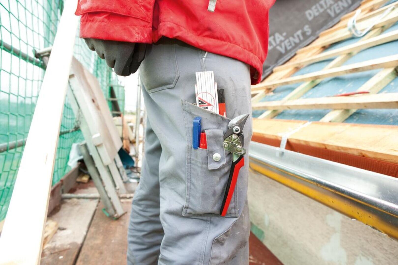 A person wearing gray work pants with a pocket full of tools, including a ruler, pliers, and pens, stands on scaffolding next to a partially constructed or repaired sloped roof.
