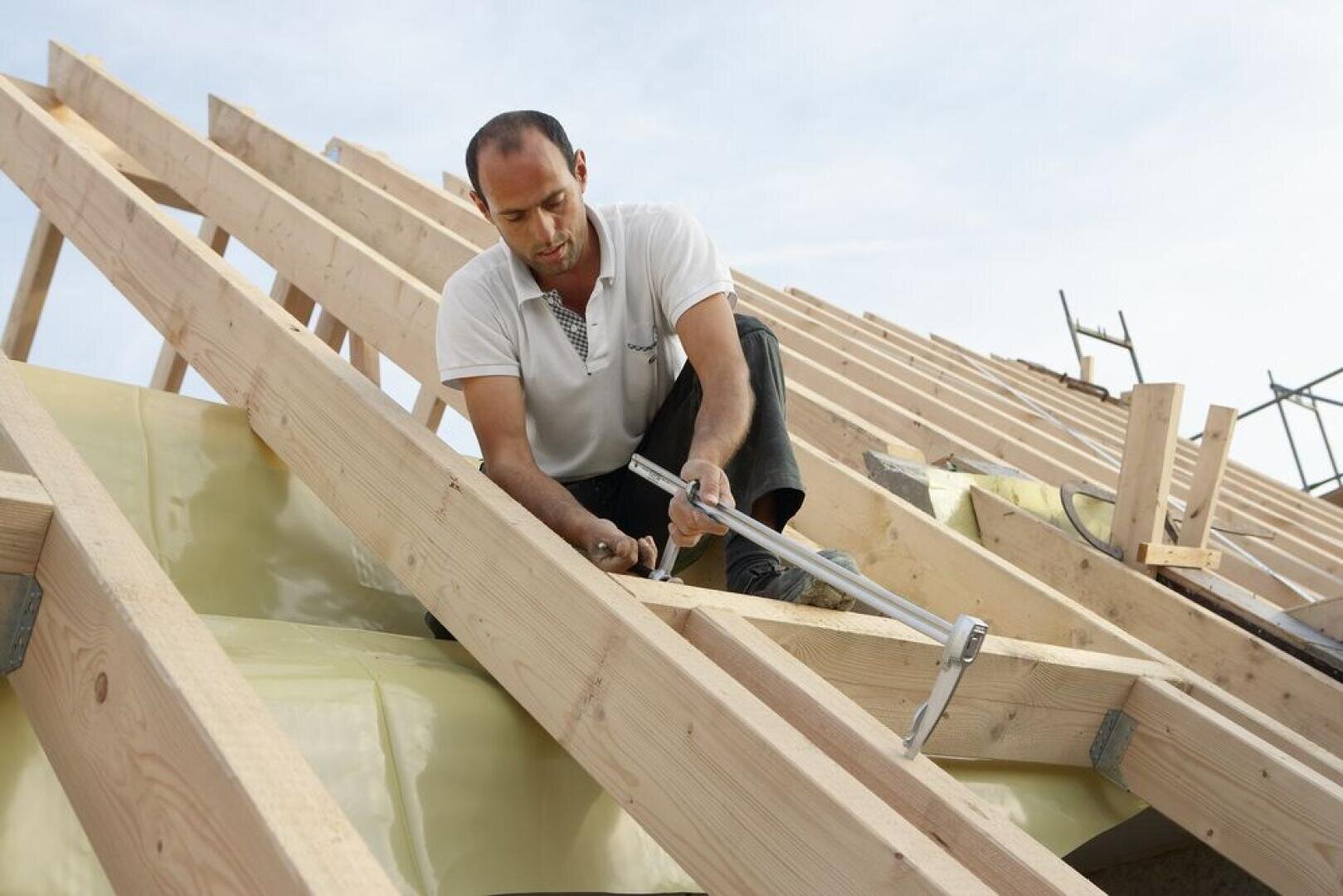 A man wearing a white shirt is kneeling on a wooden roof frame, using a hammer and nails to secure beams, with construction materials and blue sky in the background.