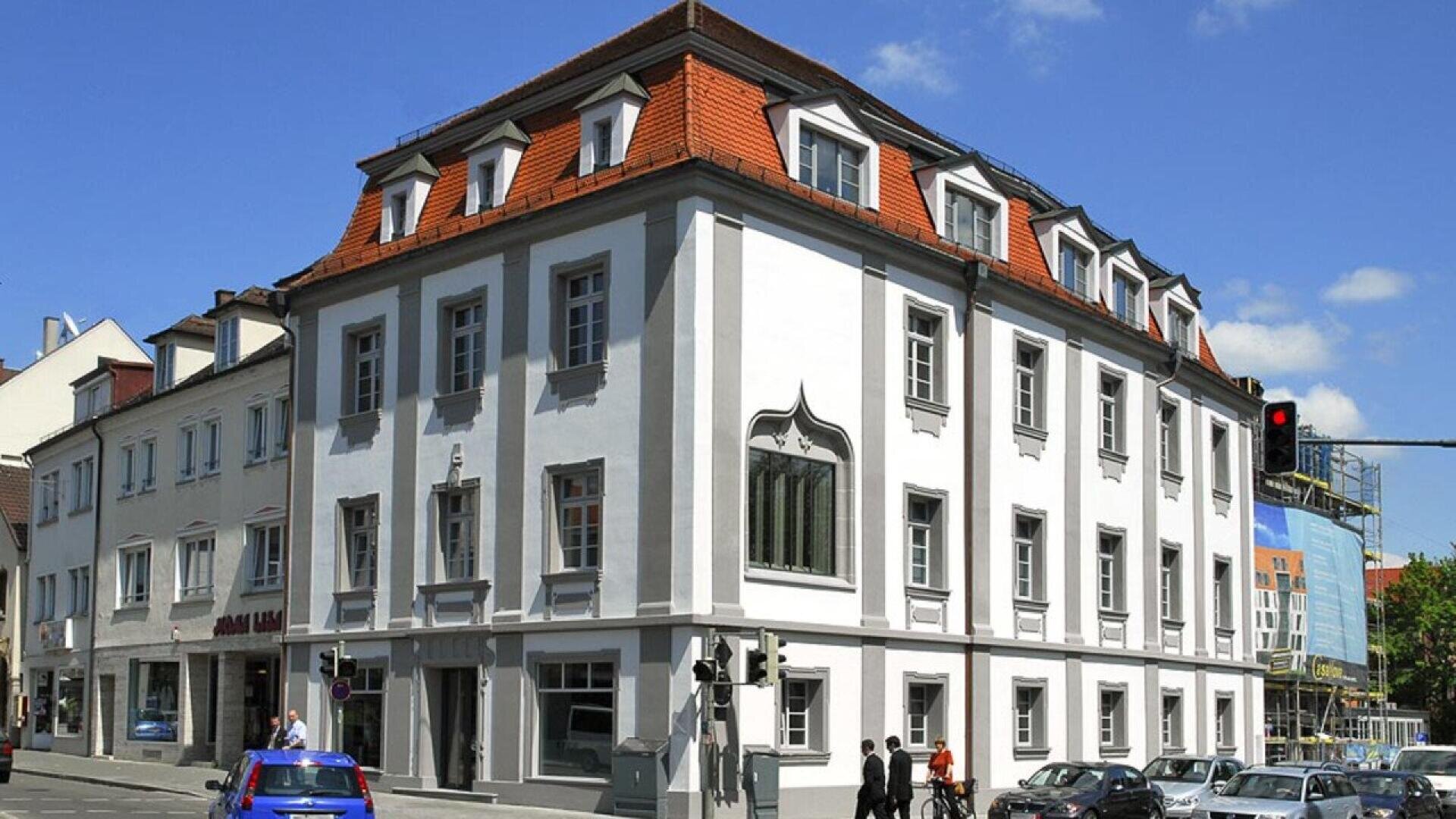 A white, three-story building with a red-tiled roof stands on a street corner. People walk along the sidewalk, and cars wait at a traffic light under a blue sky with a few clouds.