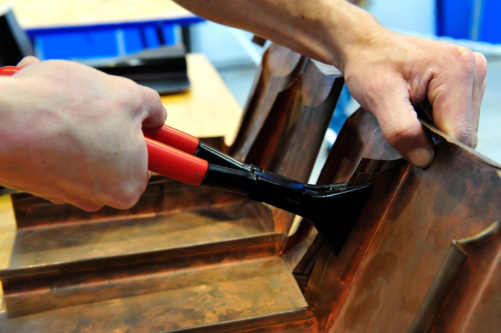 Close-up of hands using red-handled pliers to shape or bend a piece of copper sheet metal, possibly during a metalworking or crafting project.