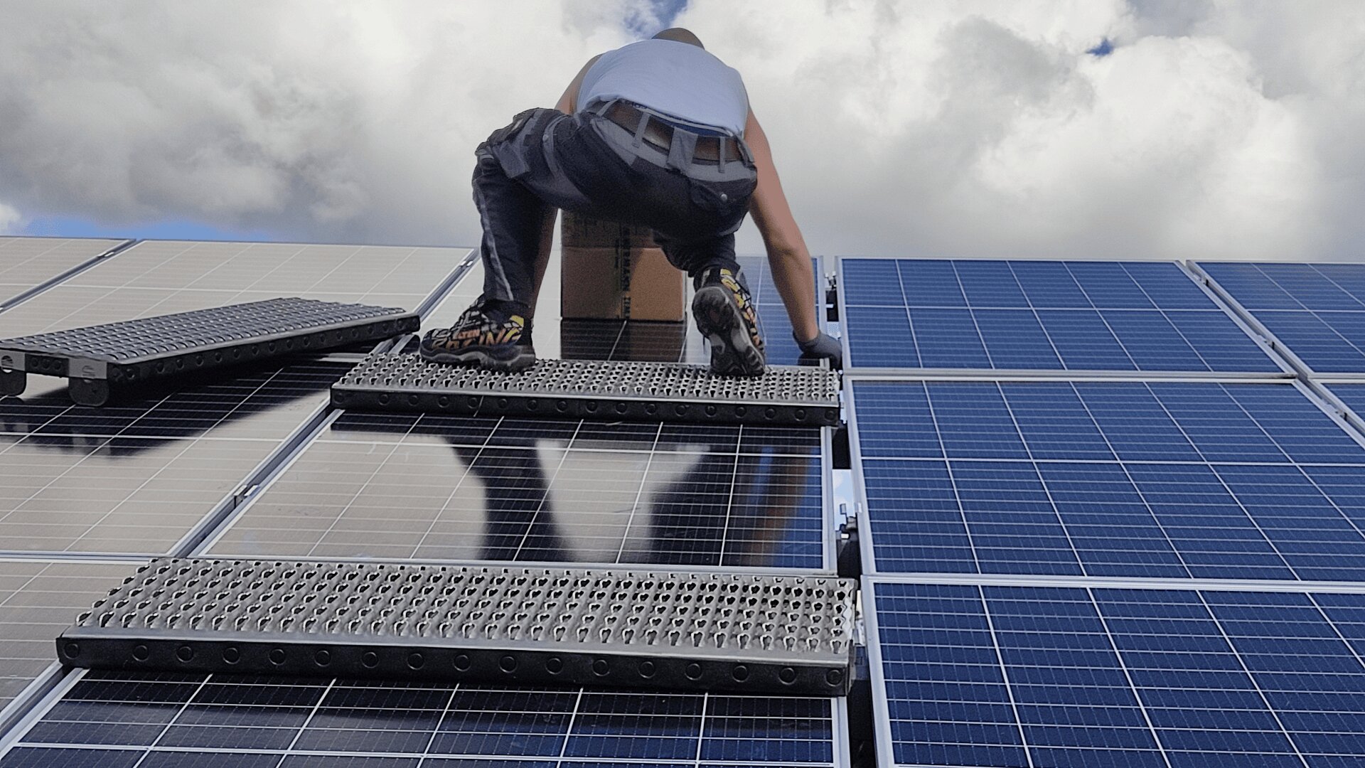 Ein Arbeiter installiert oder wartet unter einem teilweise bewölkten Himmel Solarmodule auf einem Dach und nutzt dabei Metallplattformen als Stütze.