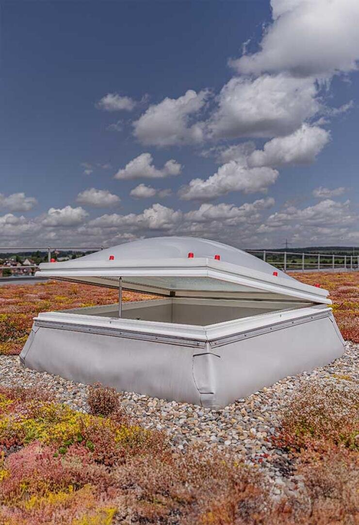 Ein weißes Dachfenster ist auf einem grünen Dach geöffnet, das mit niedrig wachsenden Pflanzen und kleinen Steinen bedeckt ist, unter einem teilweise bewölkten Himmel.