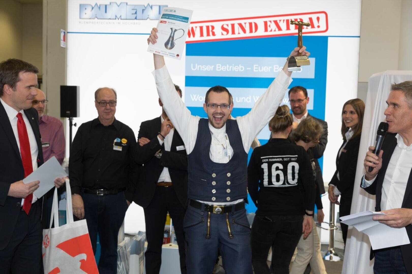 Ein Mann in traditioneller Kleidung feiert auf der Bühne, hält eine Urkunde und eine Trophäe über seinem Kopf, umgeben von lächelnden Menschen bei einer Preisverleihung. Im Hintergrund sind ein großes Schild und mehrere applaudierende Schaulustige zu sehen.