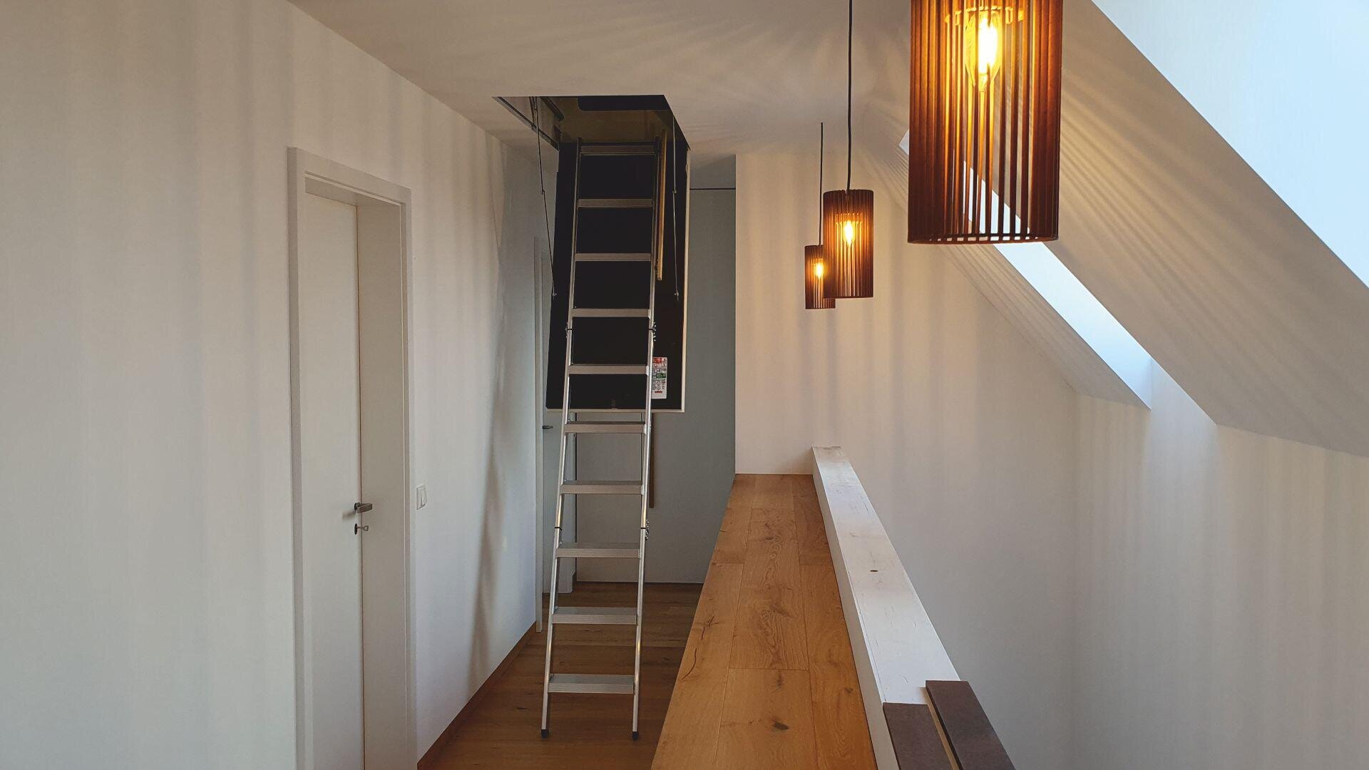 A hallway with white walls and wooden floor features a pull-down attic ladder. Three wooden pendant lights hang from the slanted ceiling, and there is a railing next to the ladder. A closed white door is on the left wall.